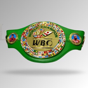 May include: A green leather boxing championship belt with a gold center plate. The center plate has the text "WBC" and "WORLD CHAMPION" with flags around the edge. Two circular medallions with boxer portraits are on each side.