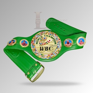 May include: A green leather World Boxing Council (WBC) championship belt. The belt features a gold center plate with the words "WORLD CHAMPION" and "WBC". Two smaller medallions with boxer portraits are on each side of the center plate. The belt has a gold buckle.