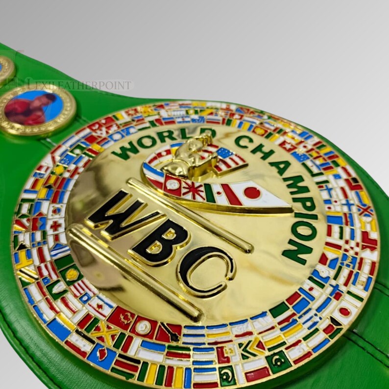 May include: A gold-colored World Boxing Council (WBC) championship belt with a green leather strap. The belt features the words "WORLD CHAMPION" and "WBC" in gold lettering, surrounded by flags of various nations.