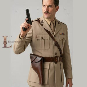 Könnte beinhalten: Eine beigefarbene Militärjacke mit einem braunen Ledergürtel und Holster-Set. Die Uniform hat eine Knopfleiste, Brusttaschen und einen Schulterriemen. Eine dunkle Handfeuerwaffe wird in der Hand gehalten. Der Text LEXILEATHERPOINT ist sichtbar.