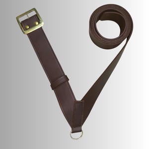 Può includere: Una cinghia in pelle marrone scuro dotata di una fibbia rettangolare in ottone con due rivetti, un passante in pelle scorrevole e un anello circolare in ottone fissato all'estremità inferiore. La cinghia è parzialmente arrotolata in alto.