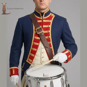 May include: A dark blue military-style jacket with red lapels, gold trim, and brass buttons, worn over a cream vest. A brown leather cross-body strap with a brass buckle is visible. The attire includes white gloves and a white snare drum.