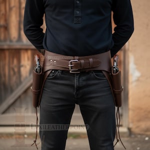 May include: A brown leather gun belt with two holsters, each holding a revolver. The belt has a silver buckle and is worn around the waist. The holsters are secured with leather ties. The image is of a person wearing the belt.