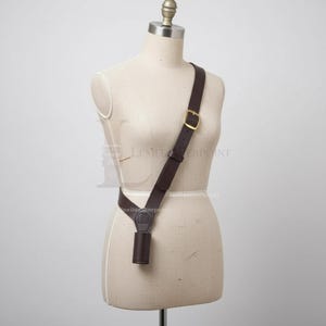 Puede incluir: Una funda de hombro de cuero marrón oscuro con una hebilla de latón y una pequeña bolsa cilíndrica. La funda se muestra en un maniquí y está diseñada para usarse cruzada sobre el cuerpo.