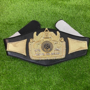 May include: A black championship belt featuring a large, ornate gold-colored central plate with an eagle emblem and the text International Boxing Council. The plate is decorated with clear stones and flanked by two smaller gold-colored side panels.