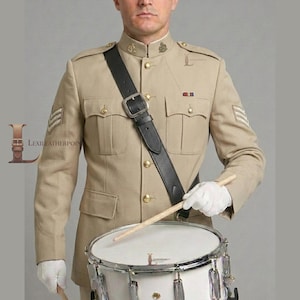 May include: A tan military-style jacket with gold buttons, chevron sleeve patches, and a black leather cross-body strap. The jacket features two chest pockets and a high collar. A white-gloved hand holds a wooden drumstick over a white drum.