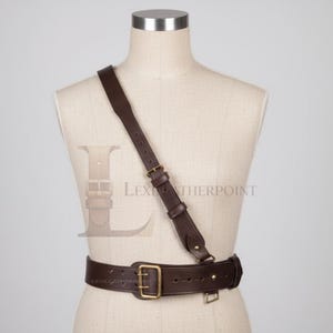 May include: A brown leather shoulder holster and belt. The holster features a shoulder strap that connects to a waist belt. The belt and shoulder strap have brass-coloured buckles. The text "LEX LEATHERPOINT" is visible on the belt.