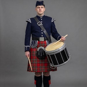 Op de afbeelding: Een muzikant draagt een donkerblauw militair jasje met zilveren details, een rood-groene tartan kilt, een zwarte sporran en een bijpassende muts. De muzikant houdt een zwart-zilveren trommel vast met twee houten drumstokken in de handen.
