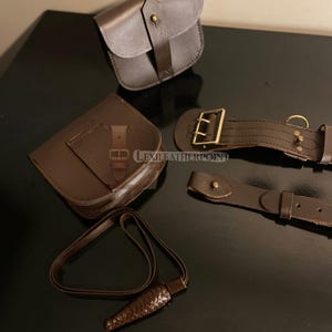 May include: A collection of brown leather accessories, including two pouches, a strap with a decorative end, and two belt-like pieces with buckles. The items are arranged on a dark surface, showcasing the texture and details of the leather.