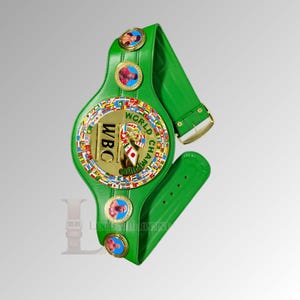 May include: A green leather World Boxing Council (WBC) championship belt. The belt features a large gold central emblem with the WBC logo and the words "World Champion". The belt has gold accents and several smaller circular emblems with portraits.