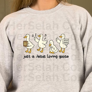 Puede incluir: Sudadera gris claro con un dise&ntilde;o de dibujos animados de cuatro gansos blancos. Un ganso sostiene un libro con la etiqueta "Holy Bible", otro canta y un tercero sostiene un letrero que dice "Jesus Saves". El texto "just a Jesus loving goose" est&aacute; debajo.