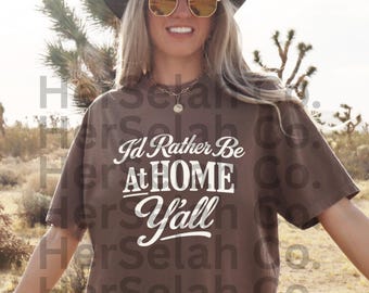 Può includere: T-shirt marrone con la scritta bianca "I'd Rather Be At Home Y'all". La maglietta è indossata con un cappello da cowboy nero e occhiali da sole dorati. Lo sfondo mostra un paesaggio desertico.
