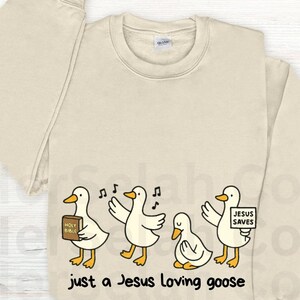 Puede incluir: Sudadera de color crema con un dise&ntilde;o de dibujos animados de cuatro gansos. Los gansos tienen picos y patas amarillas, y sostienen una Biblia, cantan notas y un letrero que dice "Jesus Saves". El texto "just a Jesus loving goose" est&aacute; impreso debajo.
