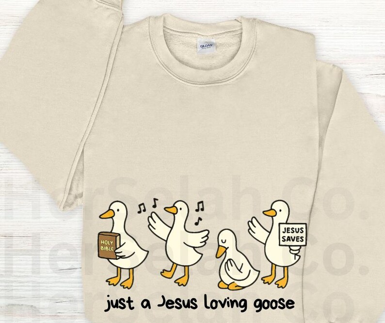 Puede incluir: Sudadera de color crema con un dise&ntilde;o de dibujos animados de cuatro gansos. Los gansos tienen picos y patas amarillas, y sostienen una Biblia, cantan notas y un letrero que dice "Jesus Saves". El texto "just a Jesus loving goose" est&aacute; impreso debajo.