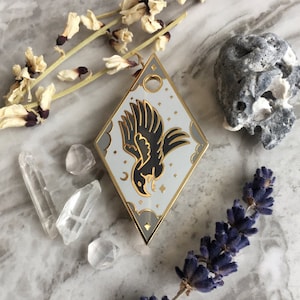 May include: A gold-toned enamel pin featuring a white background and a black silhouette of a raven flying against a backdrop of stars and clouds. The pin is shaped like a diamond.