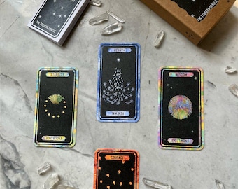 以下が含まれることがあります： 「An Oracle of Fire & Fate」というタイトルのタロットカードデッキ。茶色の箱といくつかのカードがあります。カードにはカラフルなデザインと「勇気」や「強さ」などの言葉が書かれています。透明な水晶がデッキとカードの周りに散らばっています。