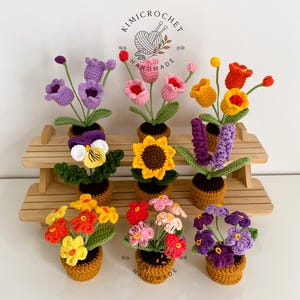 Peut inclure: Collection de fleurs crochetées à la main dans de petits pots bruns. Les fleurs sont de différentes couleurs, notamment violet, rose, jaune et rouge. L'image comprend le texte "kimicrochet" et "handmade". Les fleurs sont disposées sur une étagère en bois.