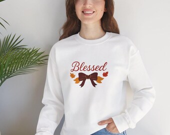 May include: White long-sleeve sweatshirt with the word "Blessed" in a script font, accompanied by a bow and autumn leaves. The design is in shades of brown, orange, and yellow. The sweatshirt is a casual, comfortable style.