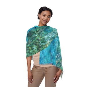 May include: A woman wearing a scarf with a water-like design in shades of blue and green. The scarf has a fringed edge and is draped over the shoulders. The scarf's colours include blue, green, and white.