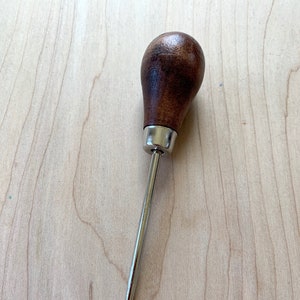 May include: A brown wooden handle with a metal awl, a tool used for piercing holes in leather.