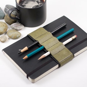 May include: A black notebook with a green leather pencil holder and three pencils. The pencils are black, blue, and white.