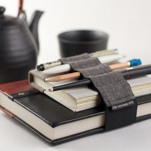 May include: A stack of two black notebooks with a gray fabric pen holder wrapped around them. The pen holder has four slots for pens and pencils. There are three pens and one pencil in the holder. The top notebook has the text "CLEVERMOMO.NET" printed on the spine.