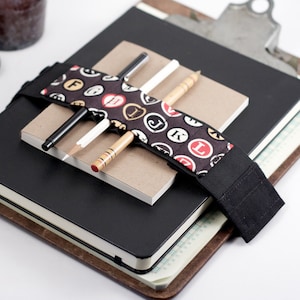 May include: A black notebook with a brown leather cover and a black elastic band with a patterned fabric insert. The band holds three writing utensils: a black pen, a white pencil, and a wooden pencil.