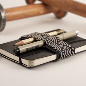 Könnte beinhalten: Ein schwarzes Notizbuch mit einem gemusterten Stoffband, das einen Bleistift und einen Stift hält.