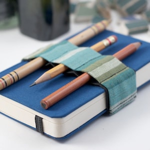 Puede incluir: Un cuaderno azul con una cubierta de tela a rayas que contiene tres lápices. Los lápices son un lápiz de madera marrón, un lápiz de madera amarillo y un lápiz rayado rojo y blanco.