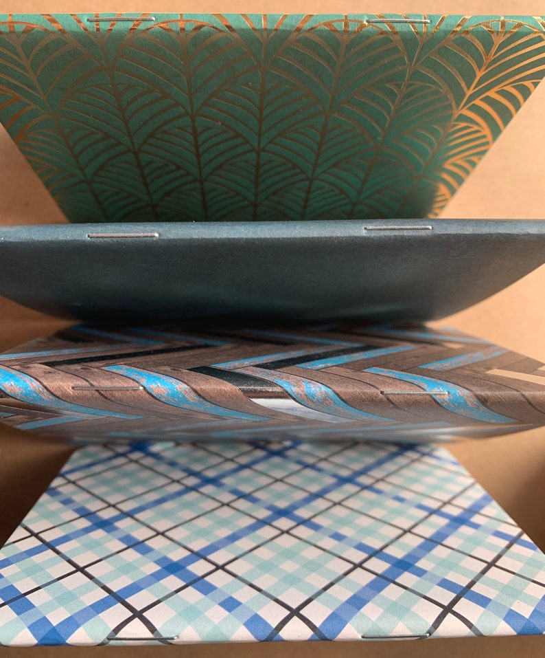 Puede incluir: Cuatro cuadernos con diferentes cubiertas estampadas. El cuaderno superior tiene una cubierta verde con un patr&oacute;n de hojas doradas. El segundo cuaderno tiene una cubierta azul oscuro. El tercer cuaderno tiene una cubierta marr&oacute;n con un patr&oacute;n de rayas azules y blancas. El cuaderno inferior tiene un patr&oacute;n de cuadros azules y blancos.