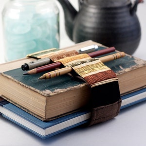 Puede incluir: Un libro vintage con una correa de cuero marrón y un marcapáginas de tela colorido que sostiene tres lápices. El libro está apilado encima de un libro azul más pequeño.