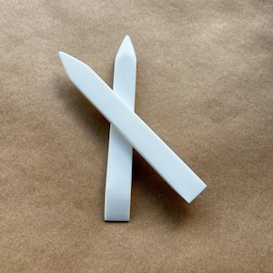 May include: Two white bone folders, essential tools for paper crafting and bookbinding, are arranged in a crossed position. The folders have a flat, rectangular body with a pointed tip, ideal for scoring and folding paper. The background is a neutral brown.