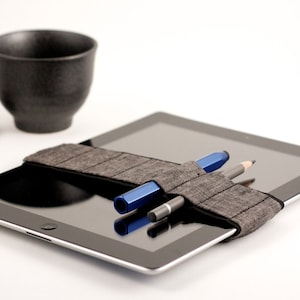 Puede incluir: Una tableta negra con una funda de tela gris que contiene dos bolígrafos y un lápiz. La tableta está sobre una superficie blanca.