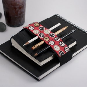 May include: A black notebook with a red and black fabric pencil holder with letters printed on it. Three pencils are in the holder. The notebook is on top of another black notebook.