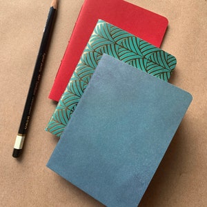 Puede incluir: Tres cuadernos apilados uno encima del otro. El cuaderno de arriba es rojo, el del medio es verde azulado con un patr&oacute;n de hojas doradas, y el de abajo es azul.
