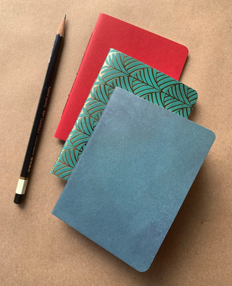 Puede incluir: Tres cuadernos apilados uno encima del otro. El cuaderno de arriba es rojo, el del medio es verde azulado con un patr&oacute;n de hojas doradas, y el de abajo es azul.