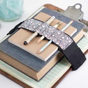 May include: A black and white patterned fabric book strap with a black elastic band. The strap is wrapped around a stack of three books and holds three pens and pencils.