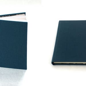May include: A dark blue hardcover journal with a white spine and a white page showing inside. The journal is closed and lying flat on a white surface.