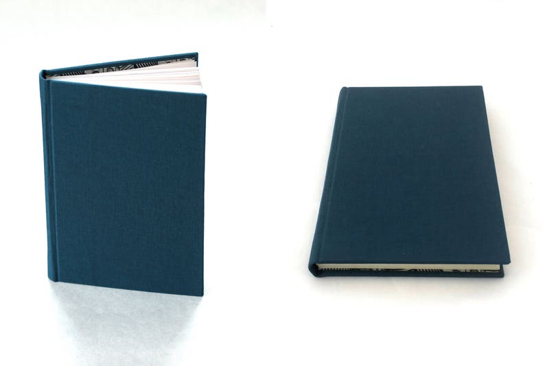 May include: A dark blue hardcover journal with a white spine and a white page showing inside. The journal is closed and lying flat on a white surface.