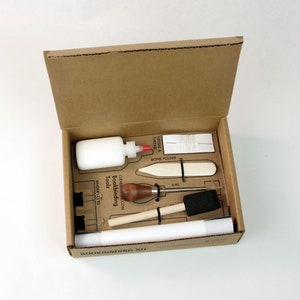 May include: A bookbinding kit in a cardboard box with tools including a bone folder, awl, binder clips, needle and thread, glue, and a wooden tool. The box has the text "Bookbinding Kit" printed on it.