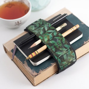 May include: A green fabric book strap with a leaf pattern wraps around a stack of two books. A black pen and two wooden pencils are held in place by the strap.
