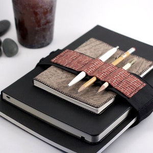 May include: Three notebooks stacked on top of each other. The top notebook has a brown leather cover and a red and white patterned pen holder strap. Three pens are inserted into the pen holder. The bottom notebook is black.