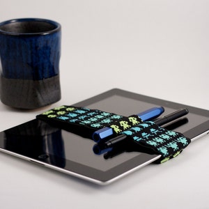 以下が含まれることがあります： 黒いタブレットコンピュータに、黒と緑の模様のゴムバンドが2本のペンを固定しています。バンドには緑色のスペースインベーダーが繰り返し描かれています。