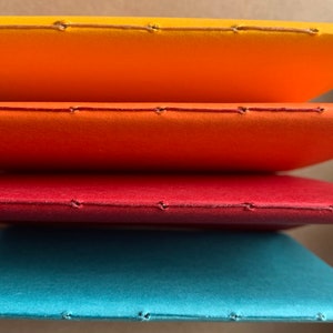 Puede incluir: Cuatro cuadernos apilados uno encima del otro. Los cuadernos est&aacute;n encuadernados con una costura a lo largo del lomo. Los cuadernos son naranja, rojo, rojo oscuro y azul verdoso.