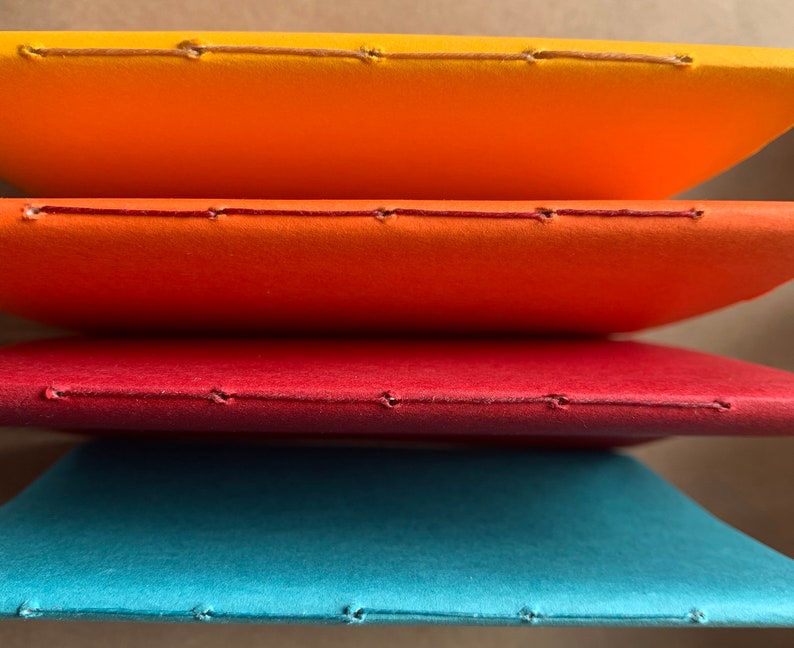 Puede incluir: Cuatro cuadernos apilados uno encima del otro. Los cuadernos est&aacute;n encuadernados con una costura a lo largo del lomo. Los cuadernos son naranja, rojo, rojo oscuro y azul verdoso.
