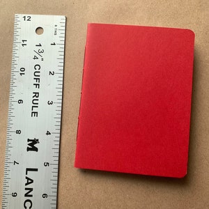 Puede incluir: Un peque&ntilde;o cuaderno de tapa dura de color rojo con una cubierta lisa. El cuaderno mide aproximadamente 10 cm de alto y 7,5 cm de ancho.