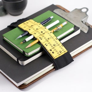 May include: A stack of three notebooks with a black elastic band holding them together. The band has a yellow measuring tape sewn onto it. There are three writing utensils in the band: a black pen, a white pen, and a yellow pencil.