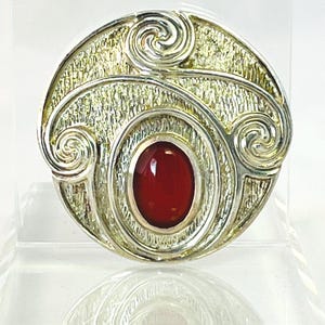 May include: A silver-toned brooch with a textured, circular design and a central, oval-shaped, red gemstone. The brooch features decorative swirls and a detailed, metallic finish, creating a vintage aesthetic.