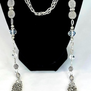May include: Silver-toned necklace with multiple strands. The necklace features decorative round beads with intricate patterns, clear crystal-like beads, and chain tassels. The necklace is displayed on a black background.