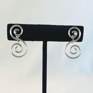 May include: A pair of silver-colored swirl earrings. Each earring features a graceful, open spiral design. The earrings are displayed on a black velvet earring stand against a soft, neutral background. The earrings are a simple, elegant accessory.